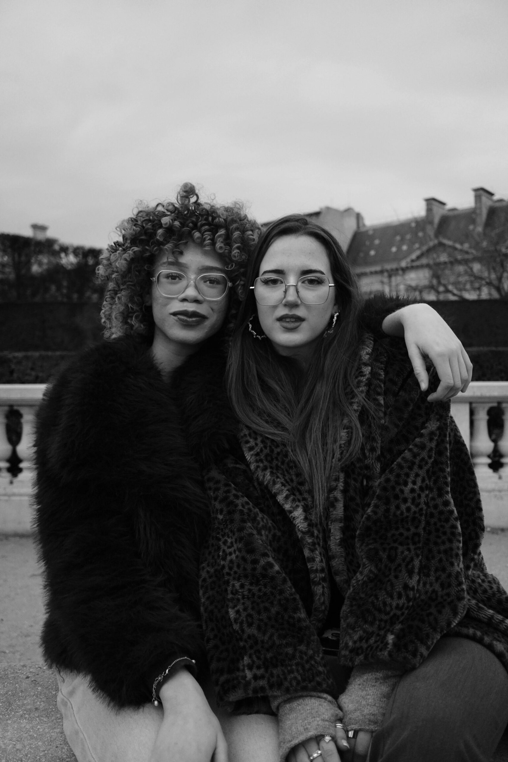 Two stylish women wearing statement faux fur coats, sitting arm-in-arm with confident expressions, representing modern wedding photographers investing in SEO and branding to attract luxury clients in 2025.