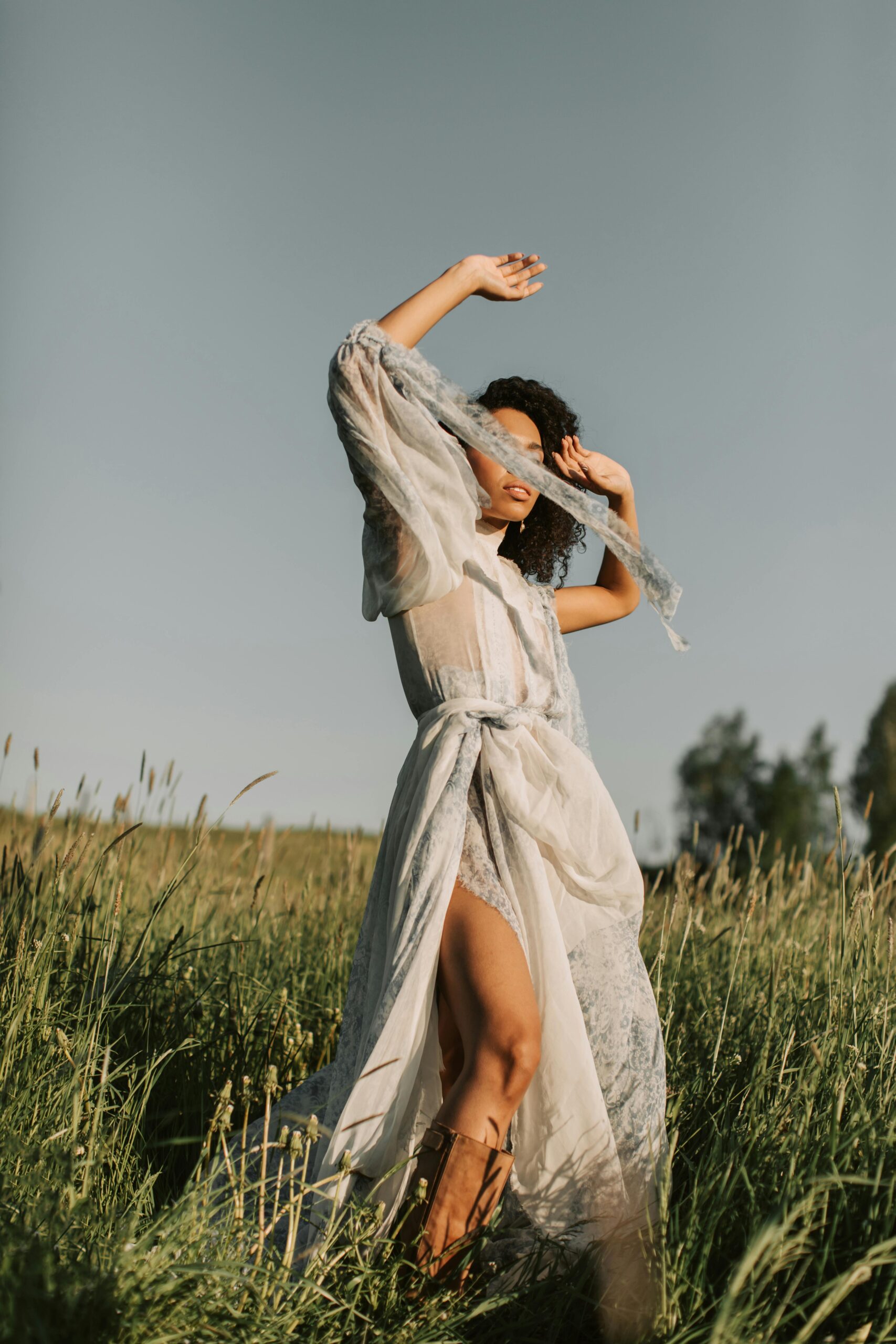 Elegant woman in a flowing vintage dress and boots stands gracefully in a sunlit grassy field, embodying creativity and inspiration for photographers during slow seasons.