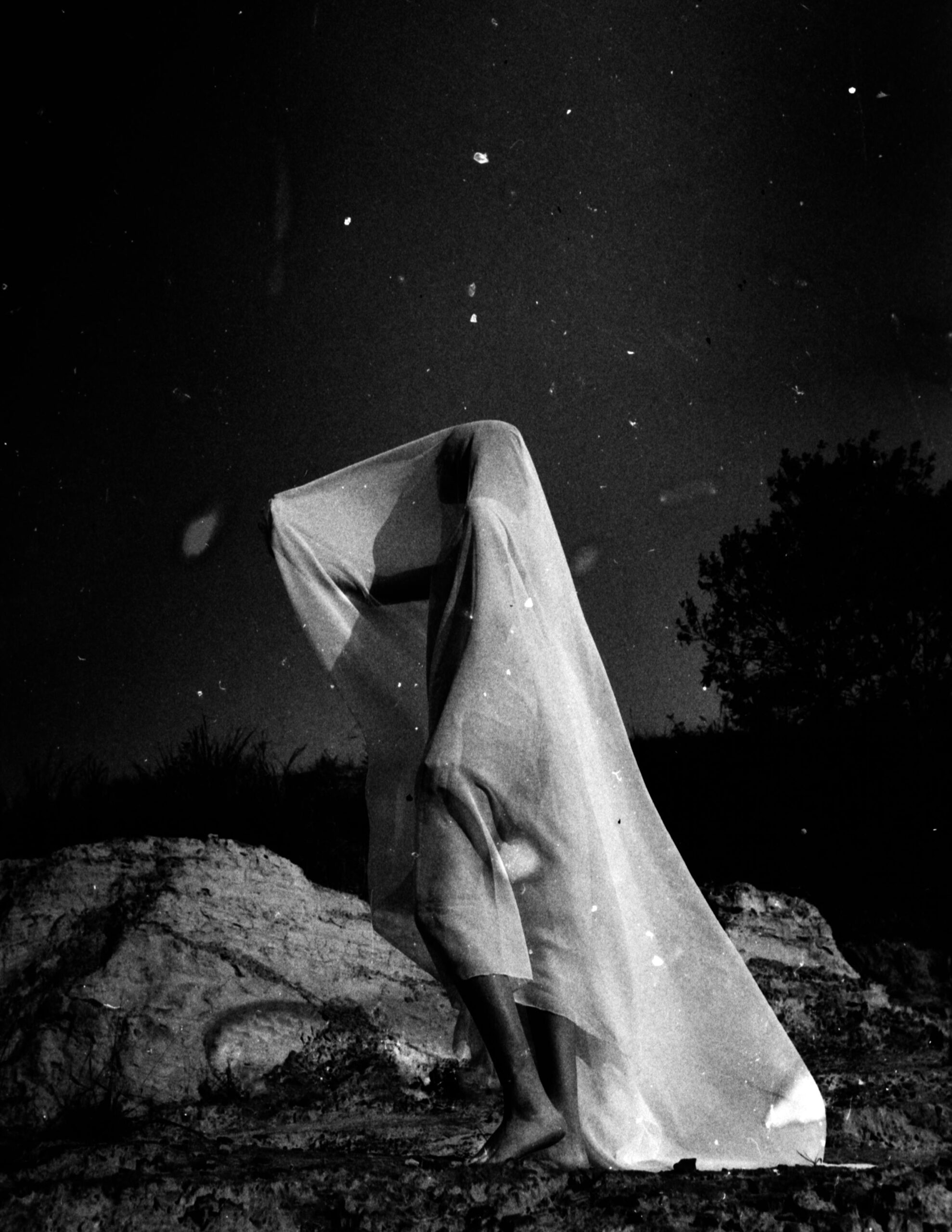 Photographer standing barefoot on rocky ground, draped in flowing fabric under the night sky—representing renewal and vision during slow season strategy work