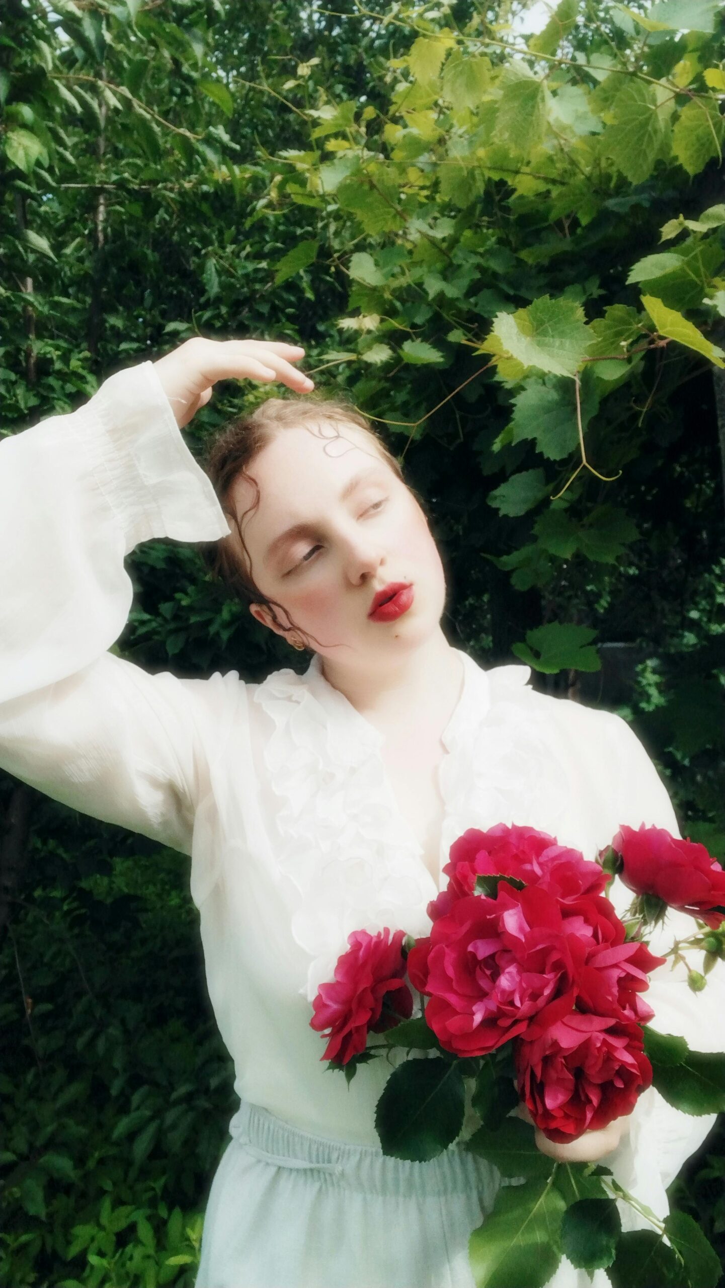 Portrait of a woman in a white vintage blouse holding vibrant red roses, surrounded by green foliage, symbolizing creativity and natural beauty in photography.