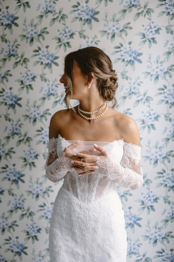 Bride in an off-the-shoulder lace gown with pearl necklace, standing against a floral wallpaper backdrop, looking to the side with hands gently clasped