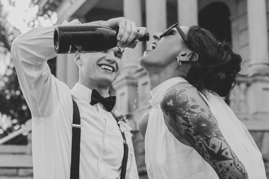 Black and white candid photo of a groom pouring champagne into the bride’s mouth while laughing together outside a historic building