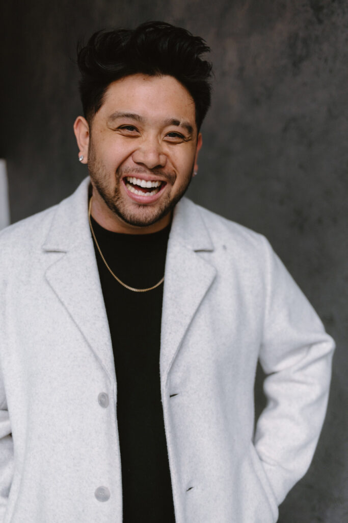 Portrait of Ean Cuenca smiling warmly while wearing a light gray coat and black shirt against a textured gray wall