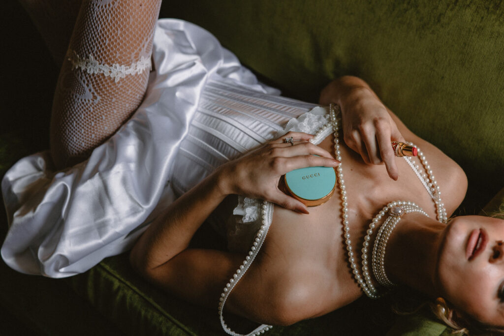 Artistic bridal portrait of a woman reclining in a white corset dress with pearl necklaces, fishnet stockings, and Gucci makeup, holding lipstick in hand