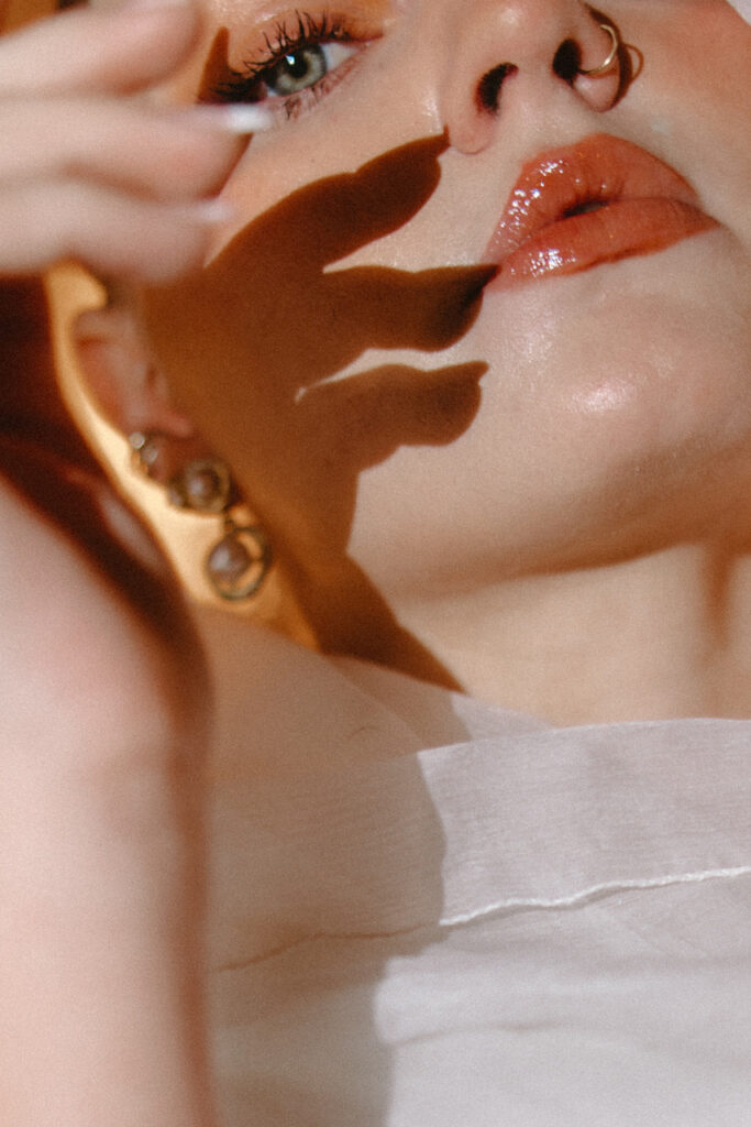 Artistic close-up of a person’s face with glossy lips, nose ring, and hand casting a shadow across the skin in warm light