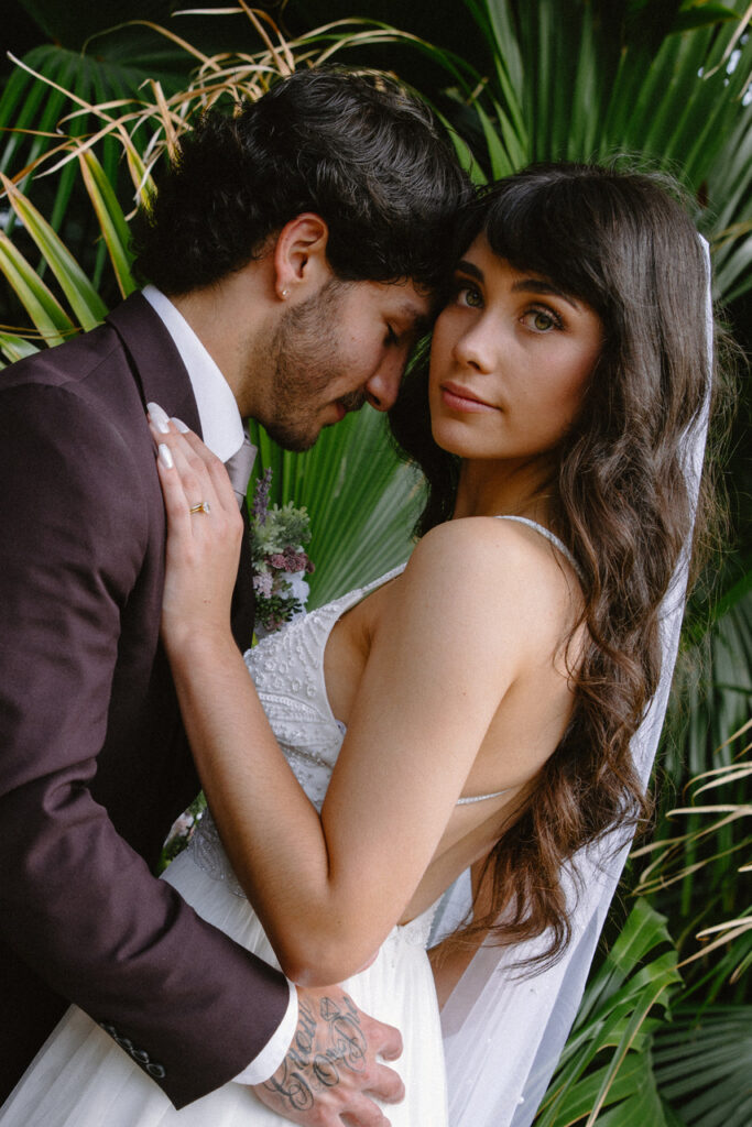 Bride and groom embracing closely in front of tropical palm leaves, with the groom leaning into the bride and the bride looking directly at the camera