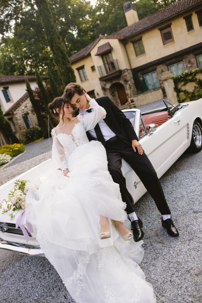 Bride and groom sitting on the hood of a white vintage convertible, leaning into each other in an intimate embrace in front of a stone villa